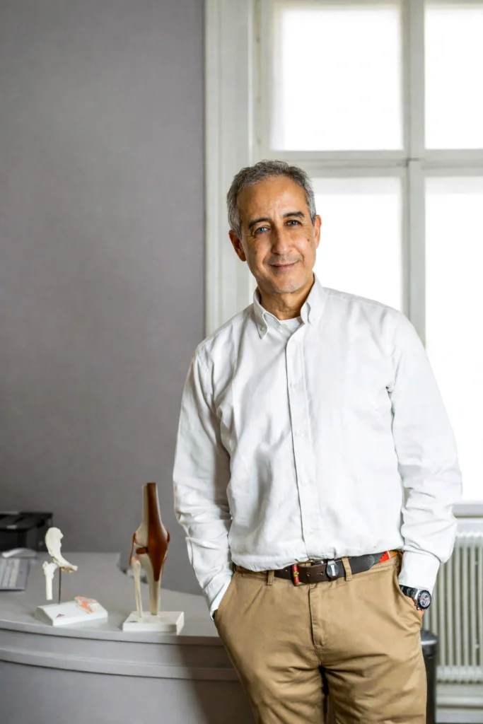 Orthopädischer Facharzt Dr. Lukas Karamat in moderner Ordination, stehend vor anatomischen Modellen des Knie- und Hüftgelenks